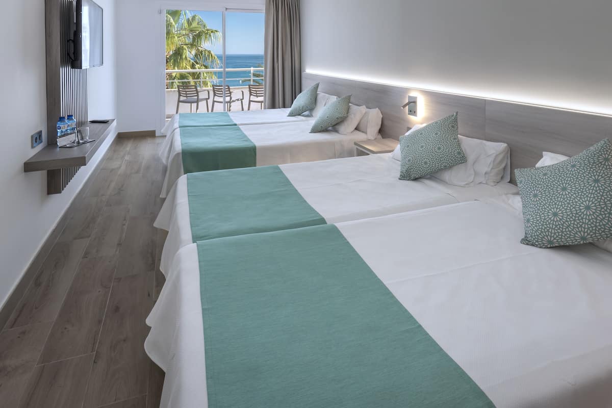 Habitación de hotel con dos camas, televisión, balcón con vista al océano y una mesa con sillas.