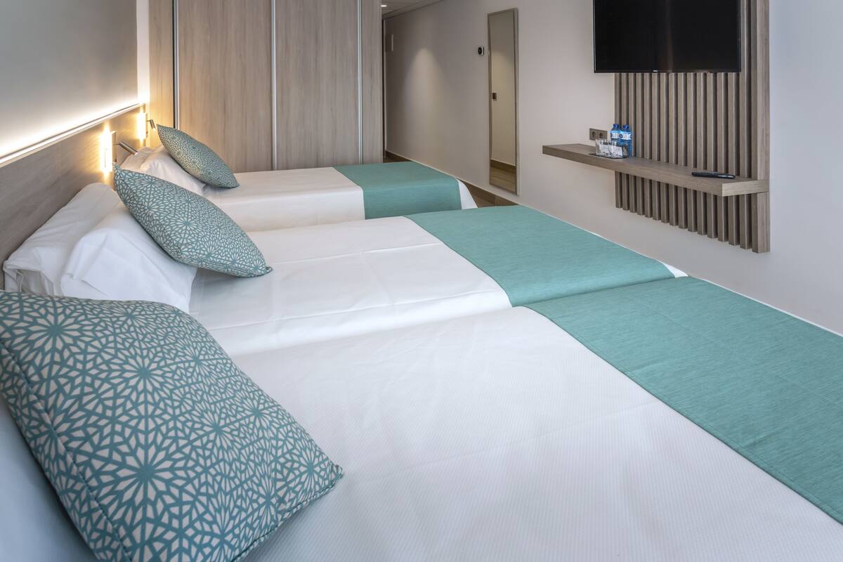 Habitación de hotel con dos camas, cada una con colcha verde aguamarina y blanca, y almohadas estampadas. Un televisor está montado en la pared, y se ve una estantería con un control remoto.