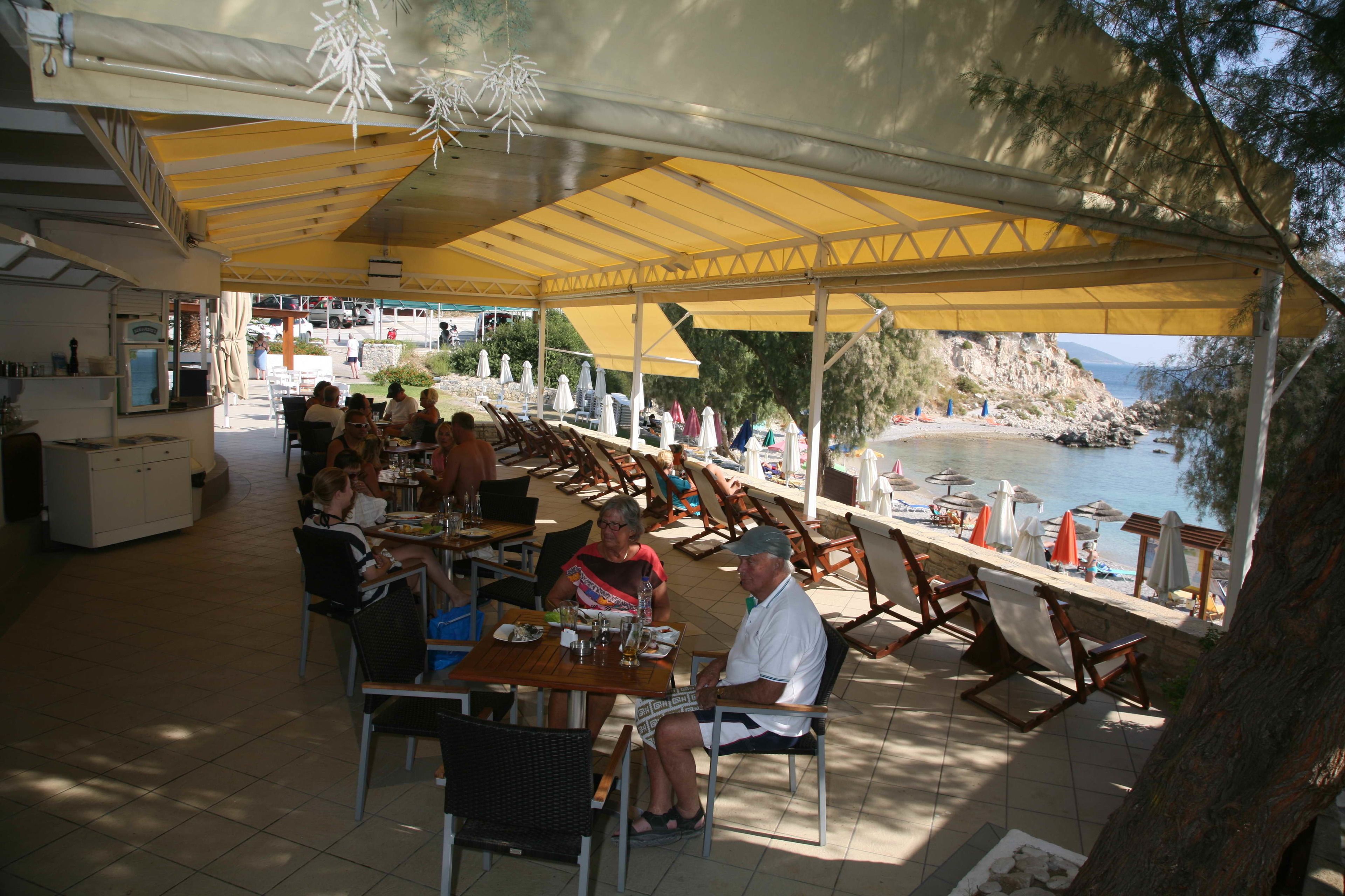 An outdoor dining area with tables and chairs under a yellow canopy, overlooking a beach and rocky coastline.