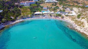 An aerial view of a coastal resort with a clear turquoise sea, a sandy beach, and several buildings with balconies overlooking the water.