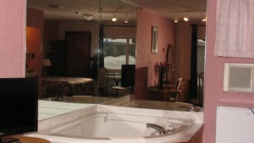 Habitación de hotel con un espejo grande, una bañera, un televisor y un comedor con una mesa y sillas.