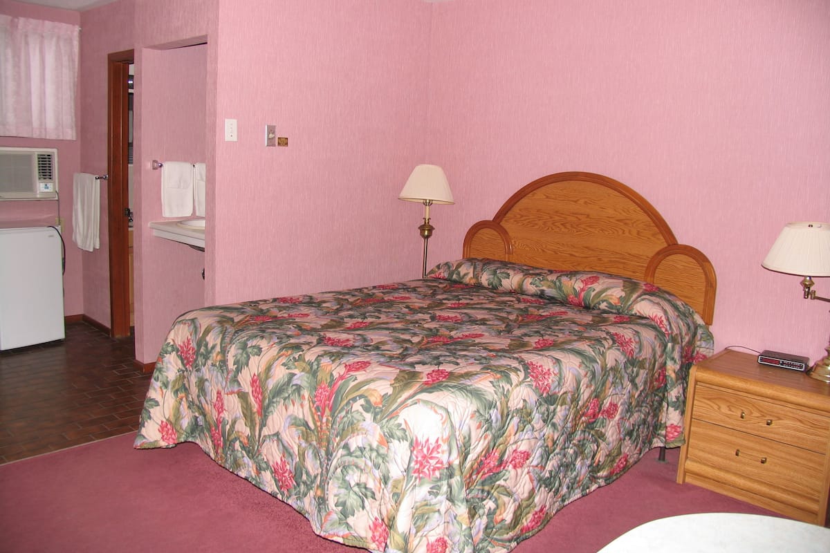 Un dormitorio con paredes rosas, una colcha de flores, un cabecero de madera, dos mesitas de noche con lámparas, una mesita auxiliar de madera y un radiador blanco.