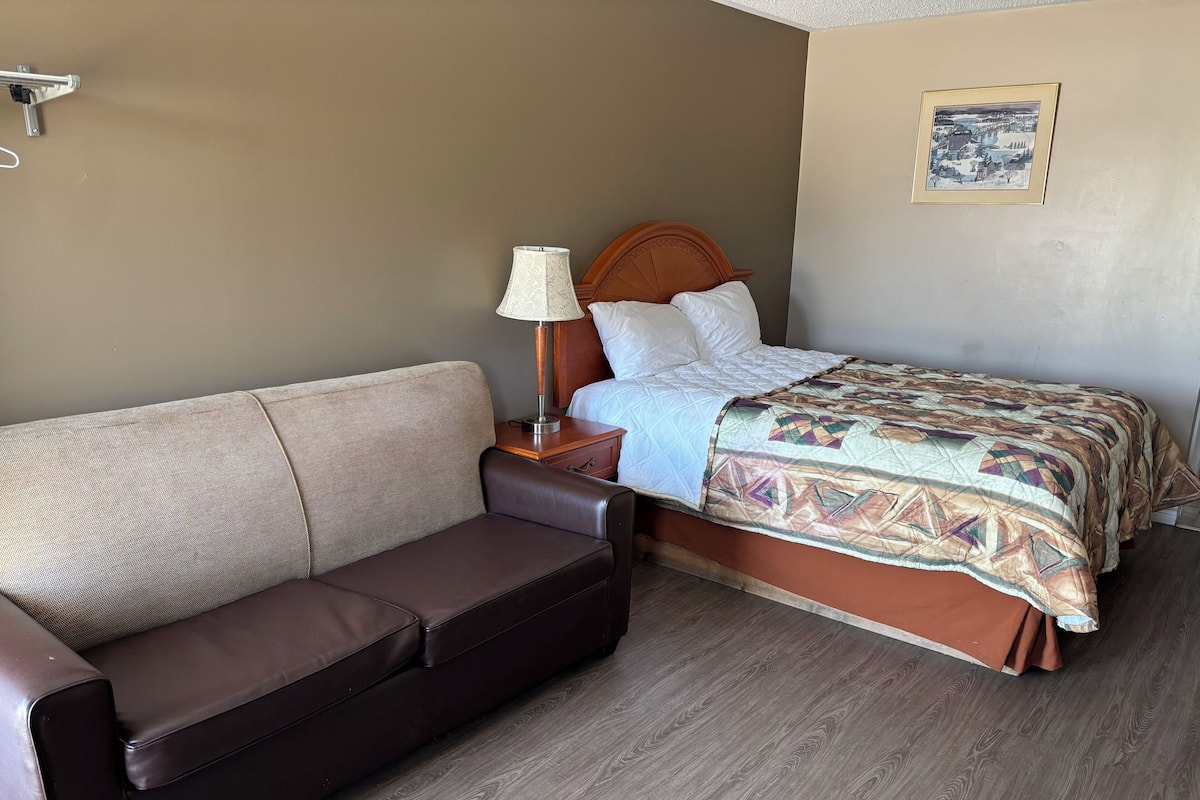 Una habitación de hotel con una cama, un sofá de cuero marrón, una mesita de noche con una lámpara y un cuadro en la pared.