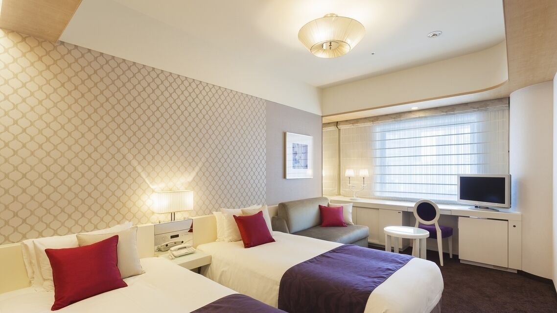 A hotel room with two beds, a desk with a TV, a small table, and a window with blinds.