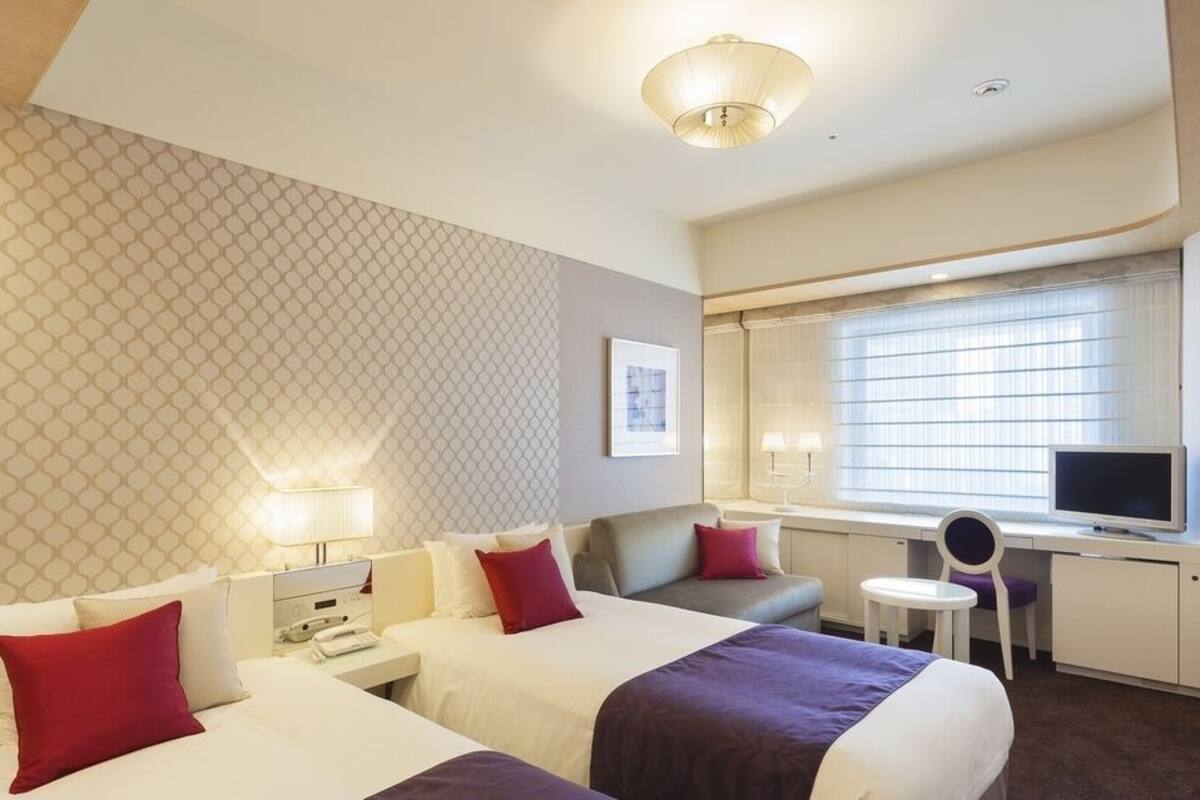 A hotel room with two beds, a desk with a TV, a small table, and a window with blinds.