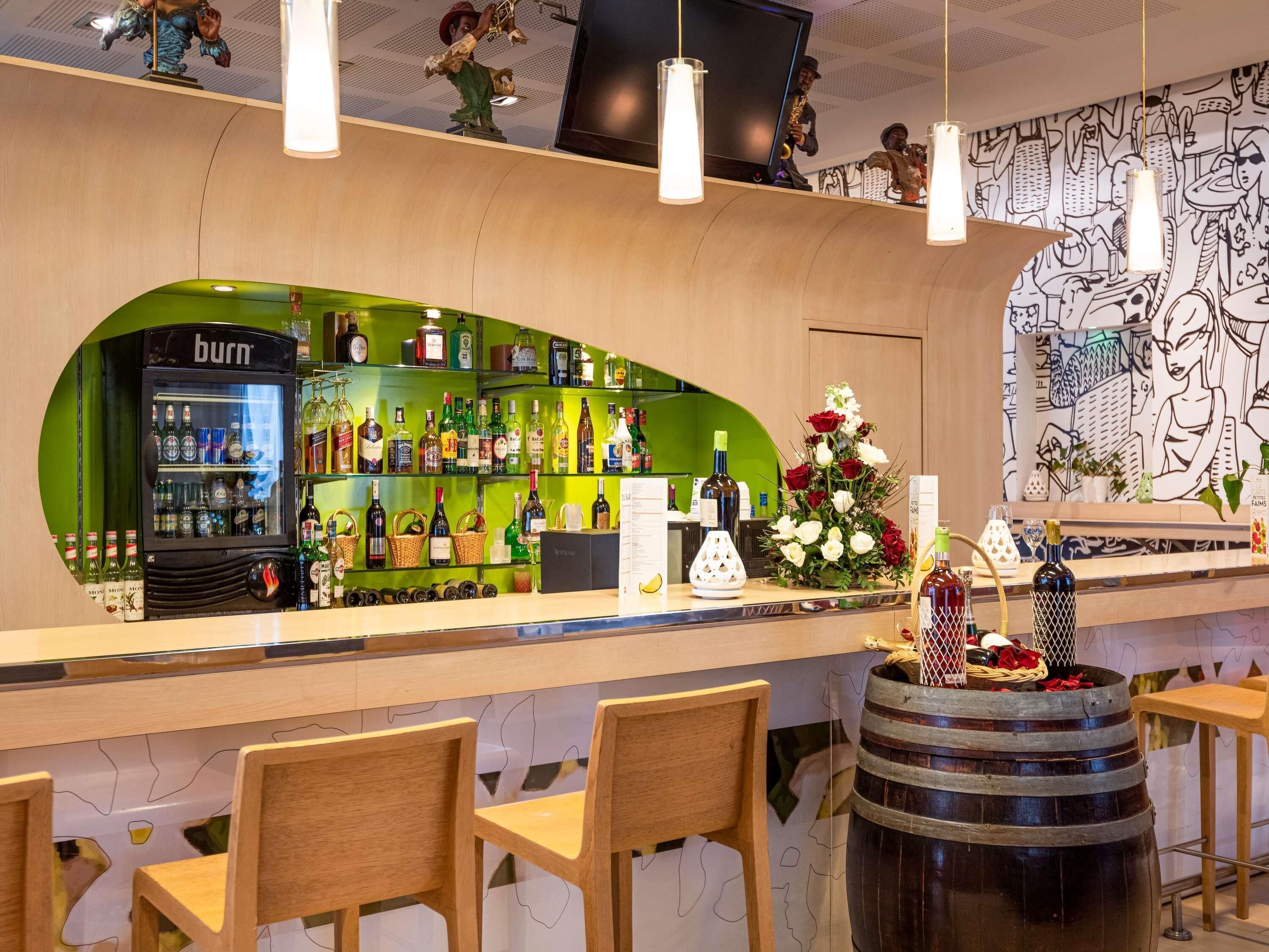 Eine Bar mit Grün im Hintergrund, Regalen voller Spirituosenflaschen, einem 'burn' Kühlschrank und einer dekorativen Blumenkomposition.