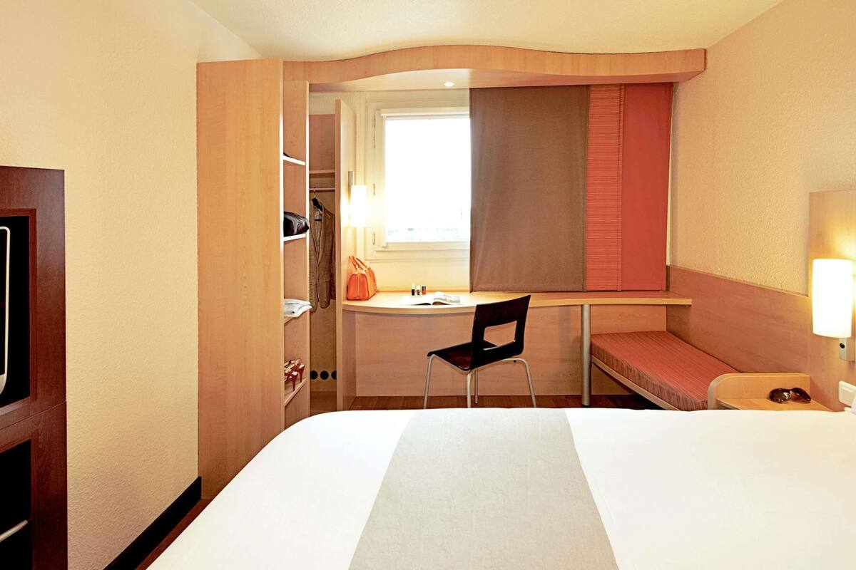 Una habitación de hotel con cama, escritorio y silla, armario y ventana con cortinas.