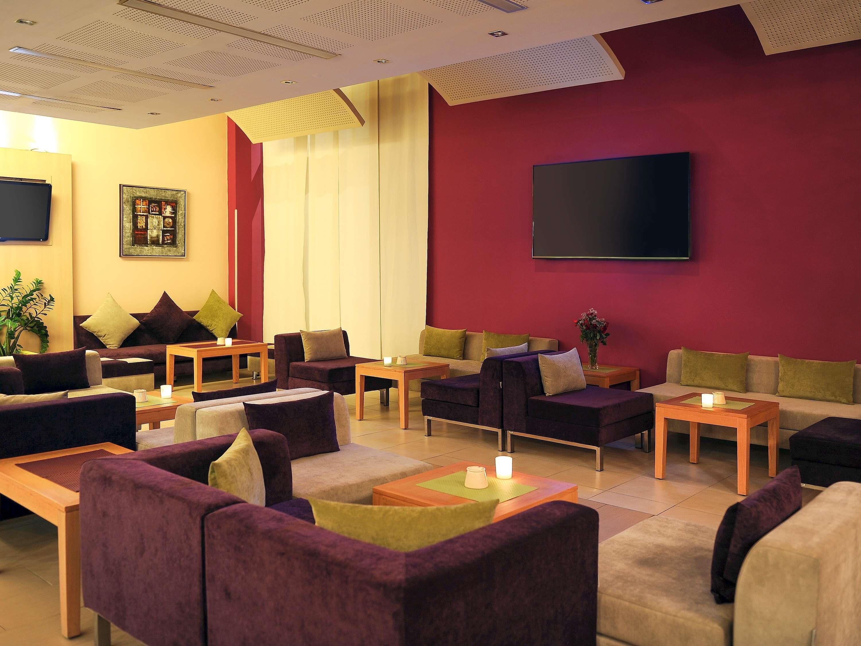 A modern hotel lobby with multiple seating areas, a flat-screen TV, and a large window with curtains.