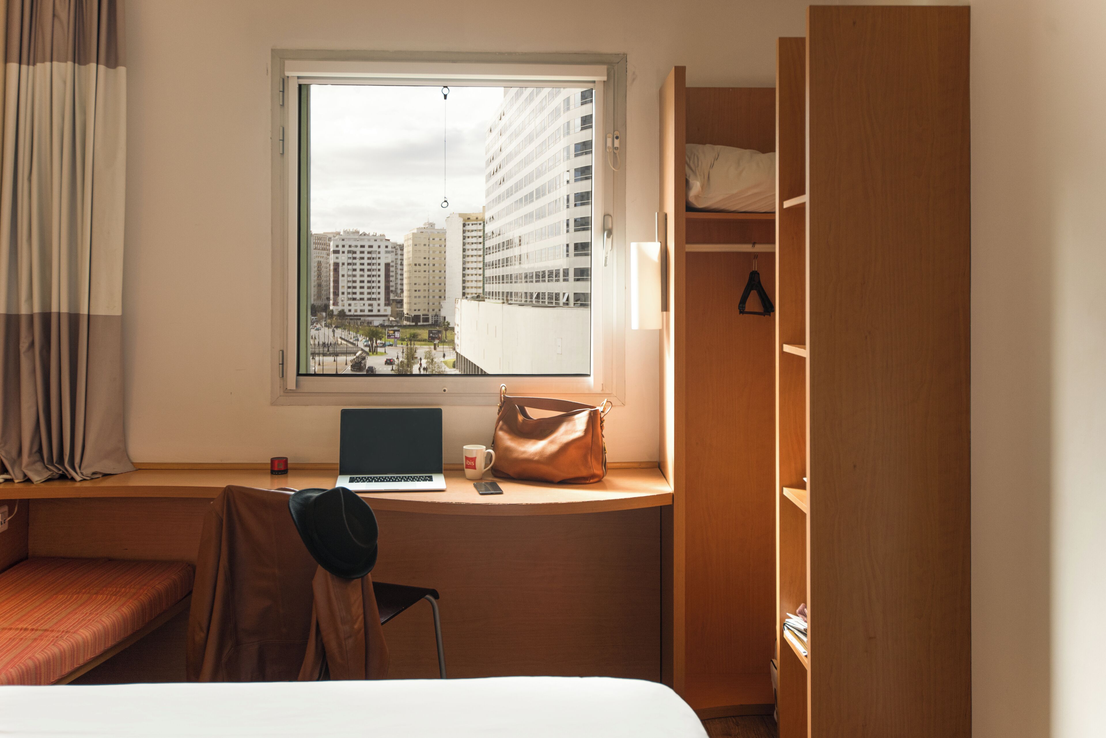 A hotel room with a bed, a desk with a laptop, a chair, a window with a city view, and a wardrobe.