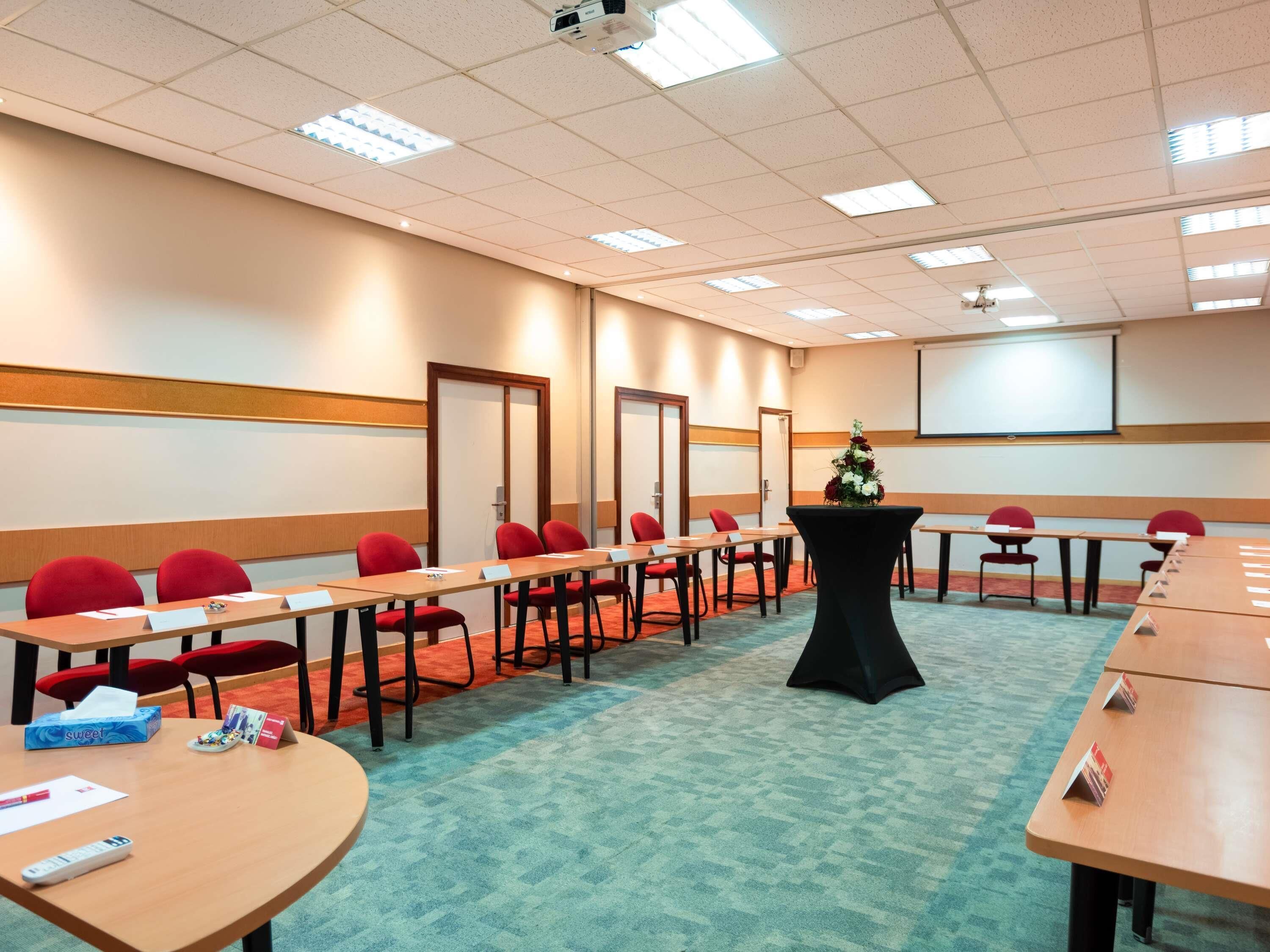 A conference room with long tables, red chairs, a projector screen, and a floral centerpiece.