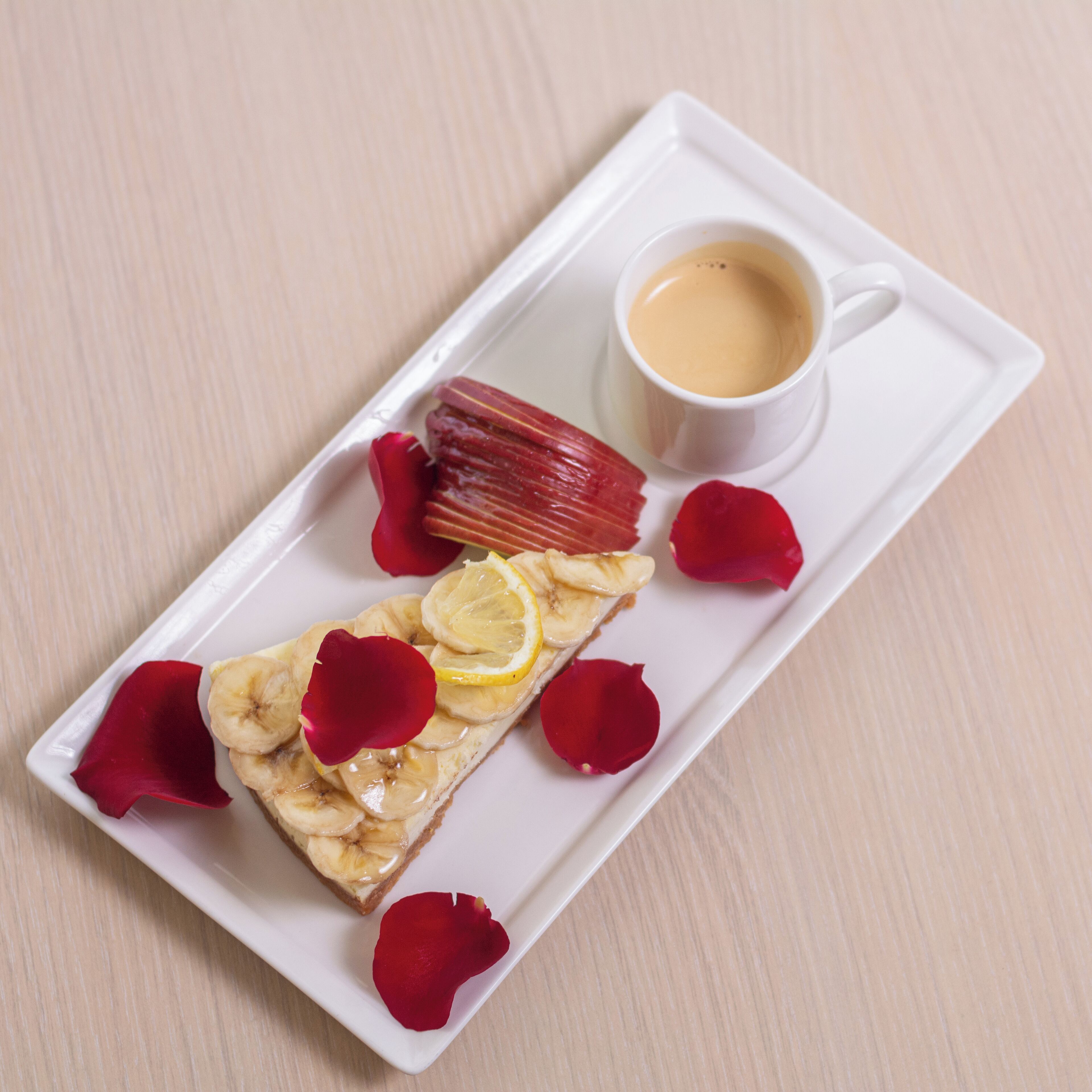 Une assiette rectangulaire blanche sur laquelle est servi un dessert garni de tranches de banane, de citron et de pétales de rose, accompagné d’une tasse de café.