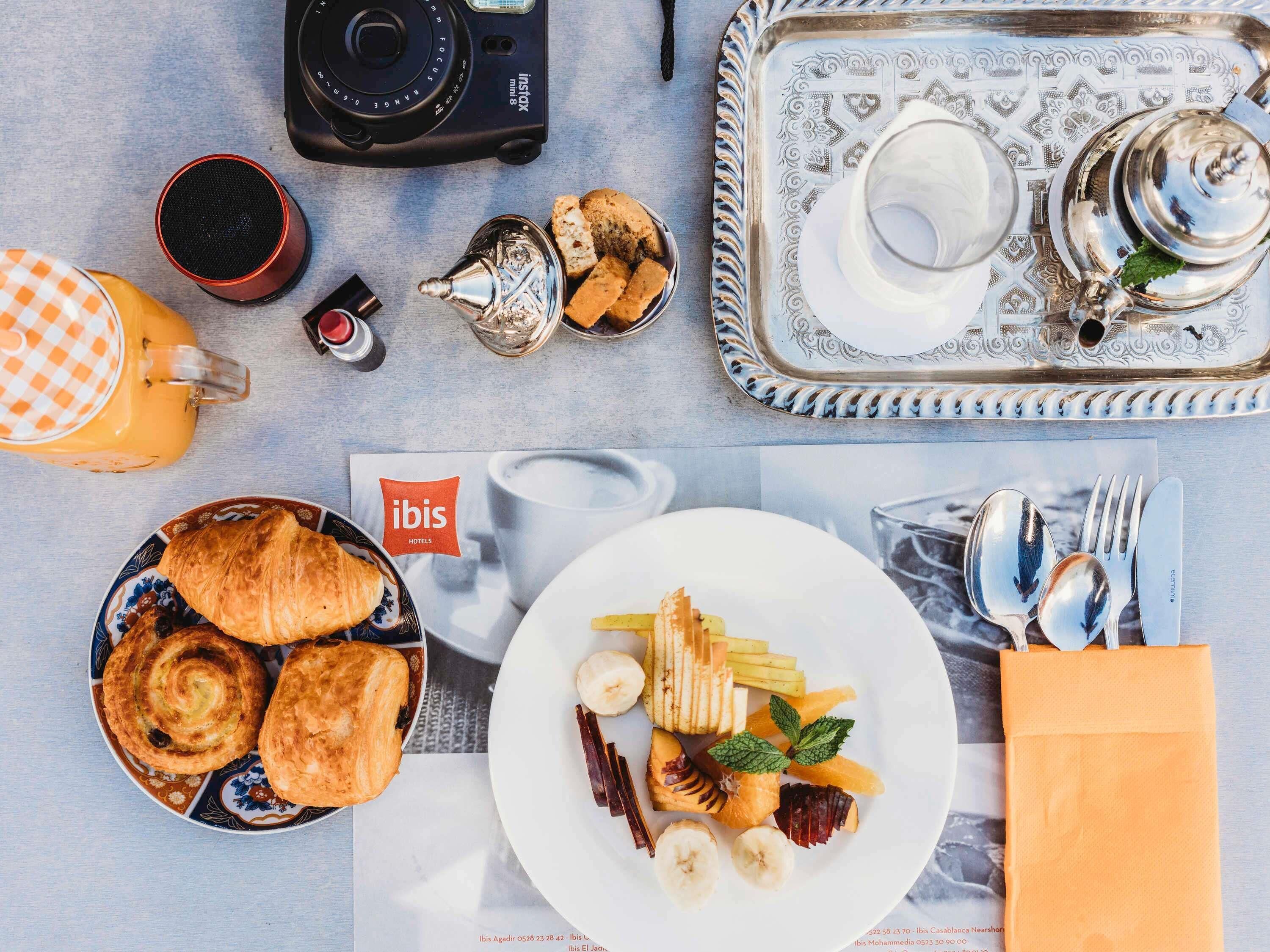 Une table dressée pour le petit-déjeuner avec des croissants, une assiette de fruits, une théière, une tasse et un appareil photo.