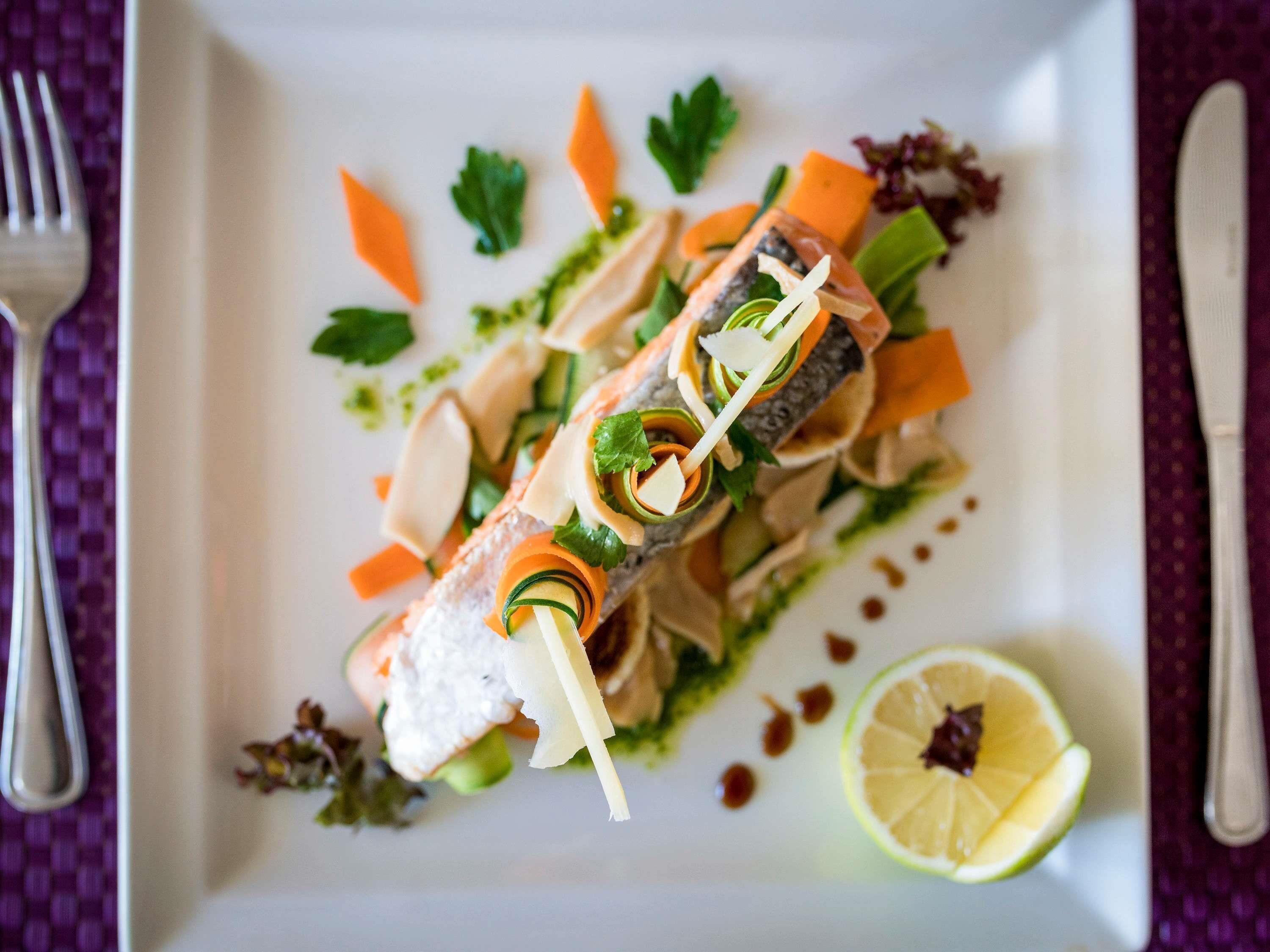 Ein weißer Teller mit Fischgericht, verziert mit Gemüse und Kräutern, dazu eine Limettenscheibe und eine Gabel.