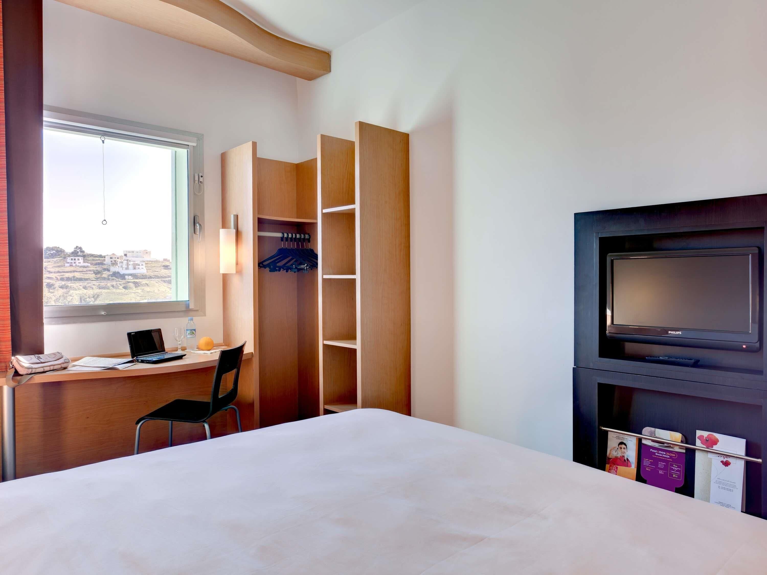 A hotel room with a bed, a desk with a laptop, a TV, and a wardrobe.