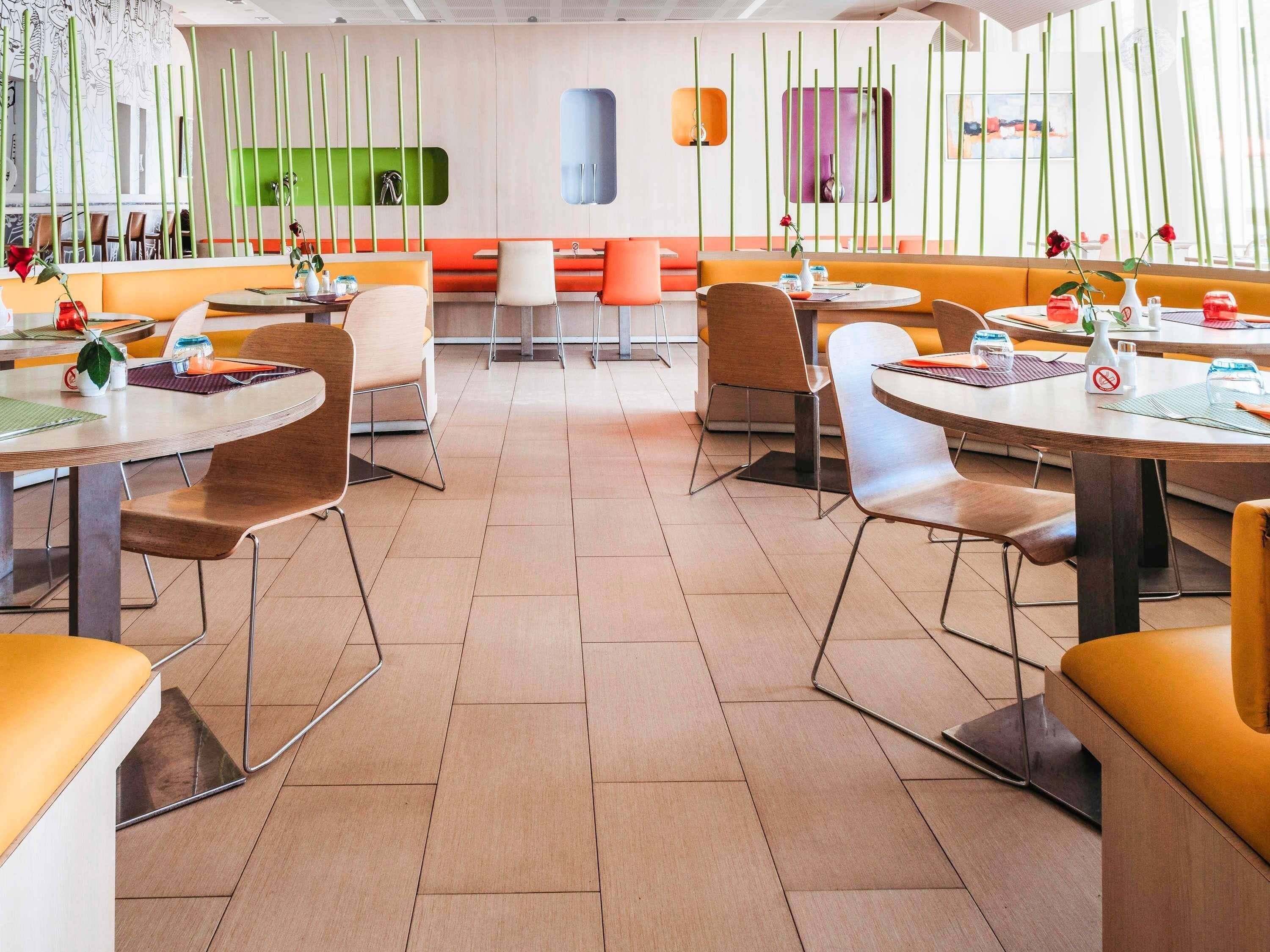 A modern restaurant with round tables and booths, a green wall with vertical lines, and orange seating.