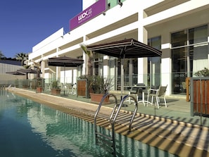 Piscina com deck de madeira e grades de metal, área de estar externa com guarda-sóis e um edifício com a placa 'WOK Restaurant'.