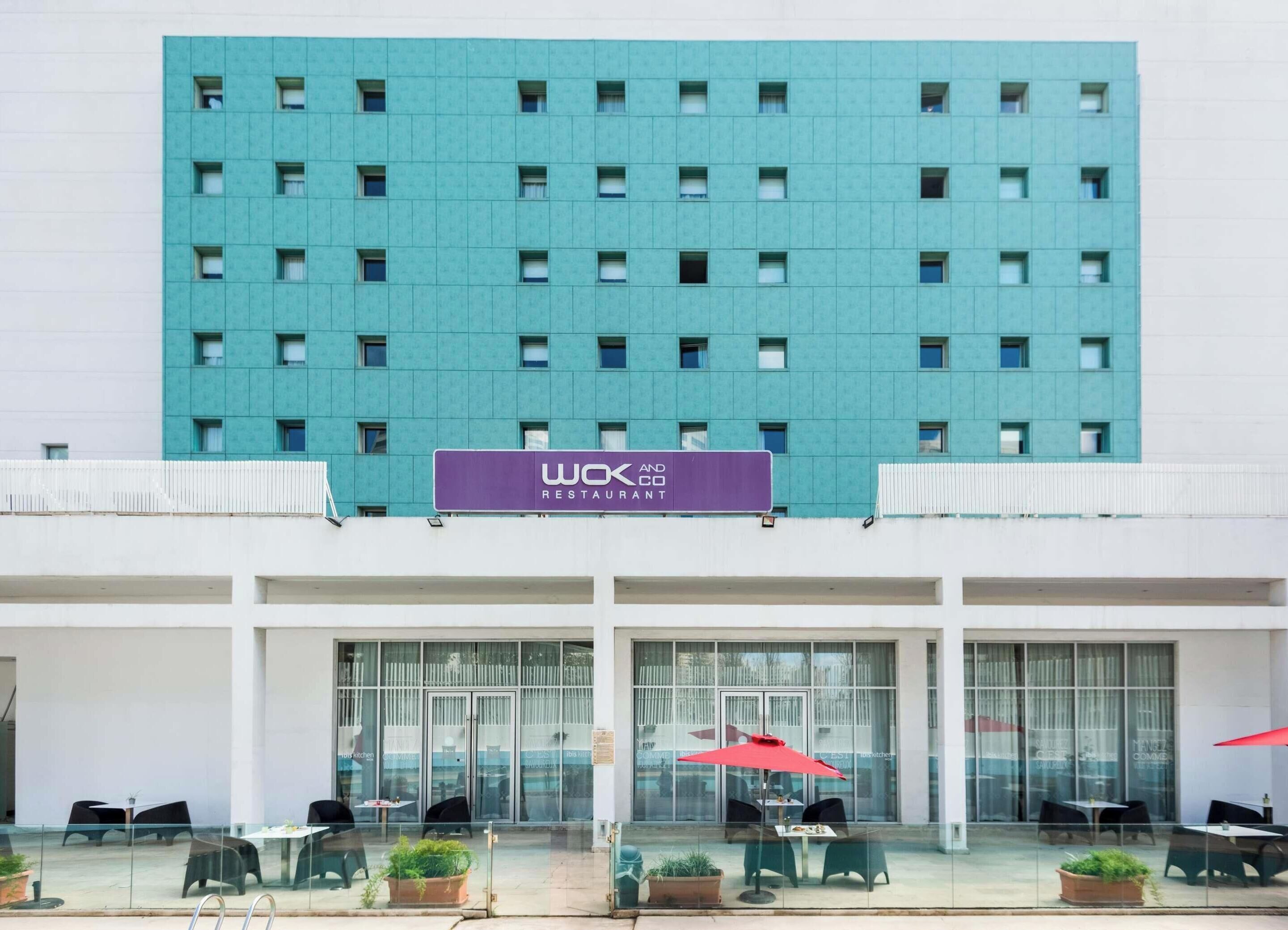 Un edificio moderno con letrero de restaurante, amplias ventanas y mesas exteriores con sombrillas.