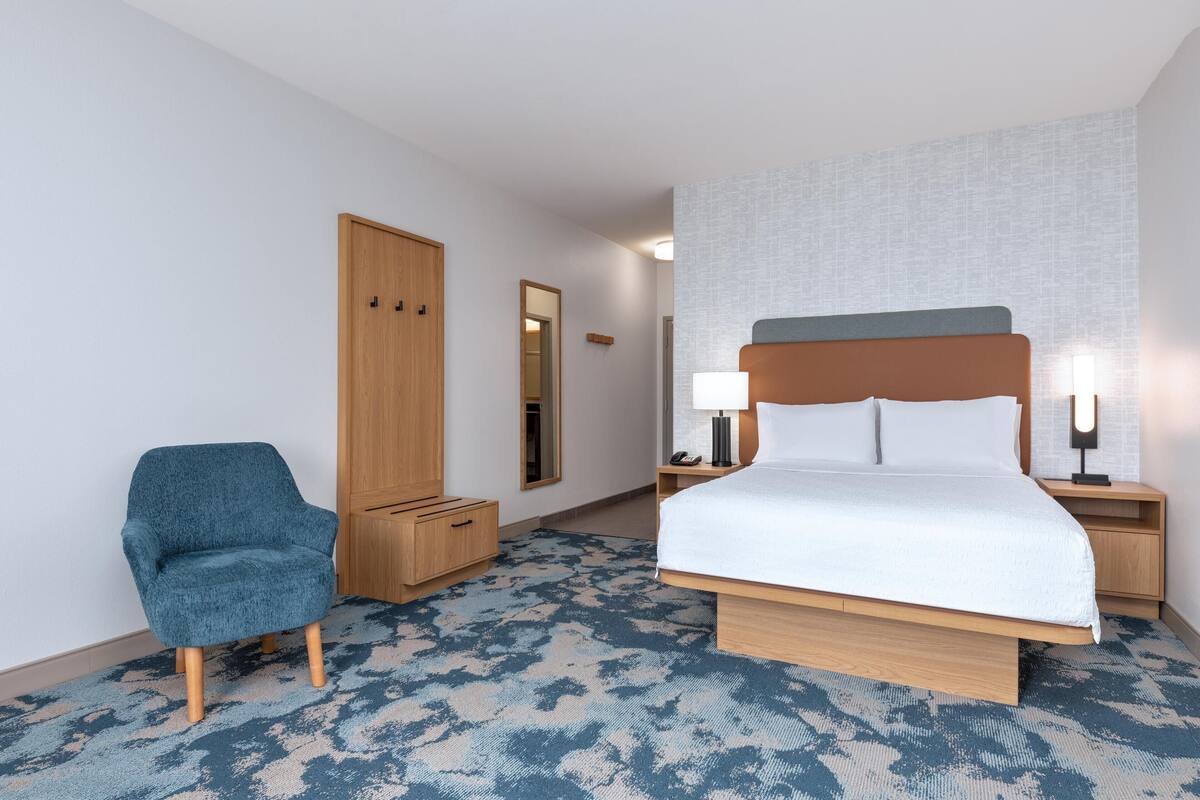 A hotel room with a bed, a chair, a wardrobe, and a nightstand with a lamp.