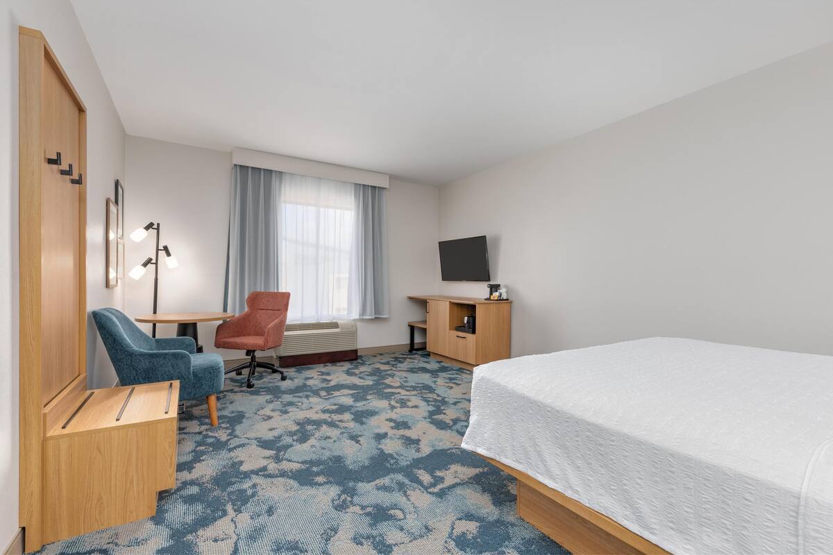 A hotel room with a bed, a desk with a chair, a television, and a window with curtains.