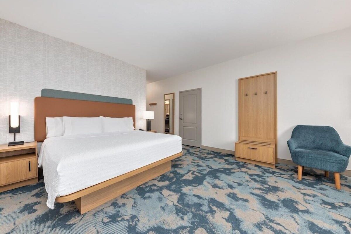 A hotel room with a bed, bedside tables, a wardrobe, a chair, and a patterned carpet.