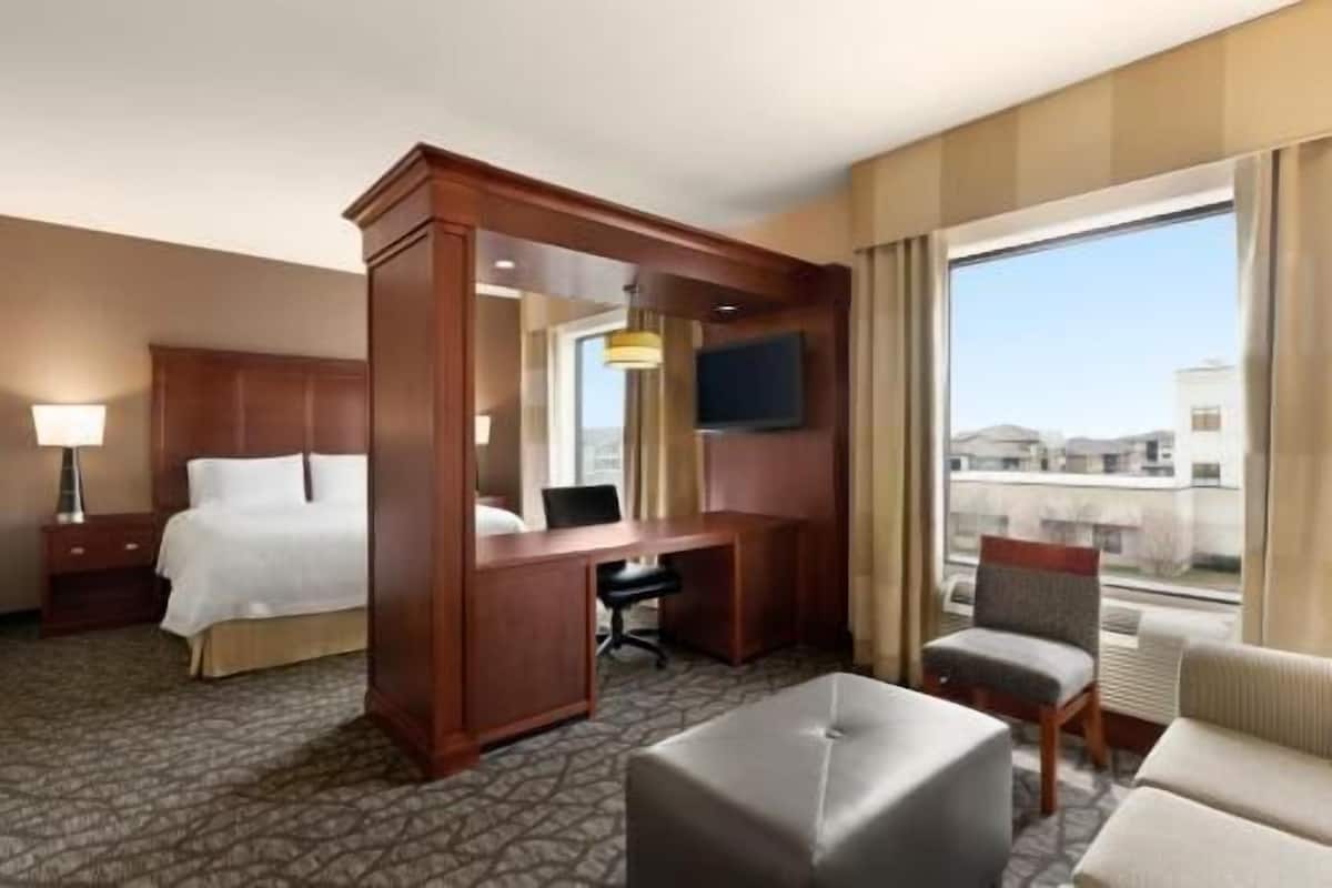 A hotel room with a bed, desk, chair, ottoman, and a large window with a view.