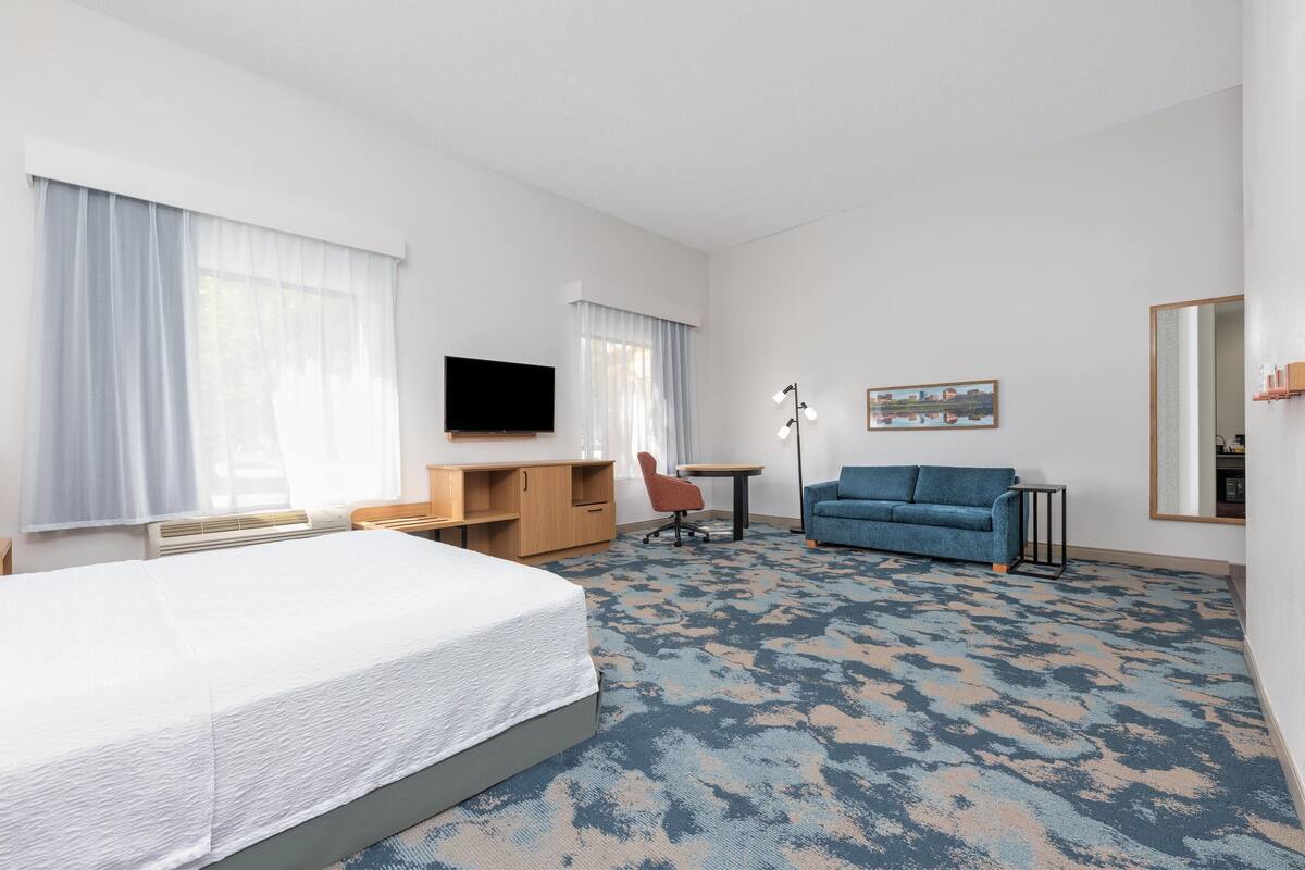 A hotel room with a large bed, a desk with a chair, a sofa, a TV, and a carpeted floor.