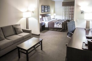 A hotel room with a sofa, a small table, a bed with a headboard, a desk with a chair, and a window with curtains.
