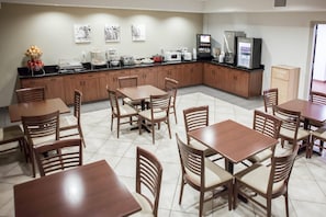 Un comedor de hotel con mesas y sillas de madera, una encimera de cocina con electrodomésticos y obras de arte en la pared.