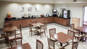 Uma sala de jantar de hotel com mesas e cadeiras de madeira, um buffet com comida e bebidas, e eletrodomésticos de cozinha.
