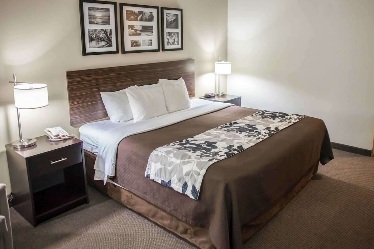 Une chambre d’hôtel avec un grand lit, deux tables de chevet, une petite table et une lampe. Des tableaux encadrés sont accrochés au mur au-dessus du lit.