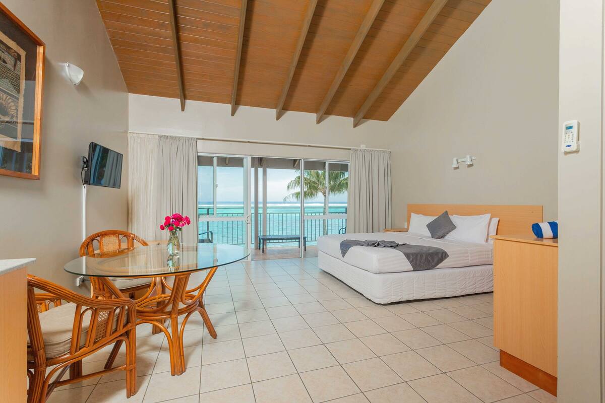 A room with a bed, a dining table with chairs, a small desk, a window with curtains, and a view of the sea.