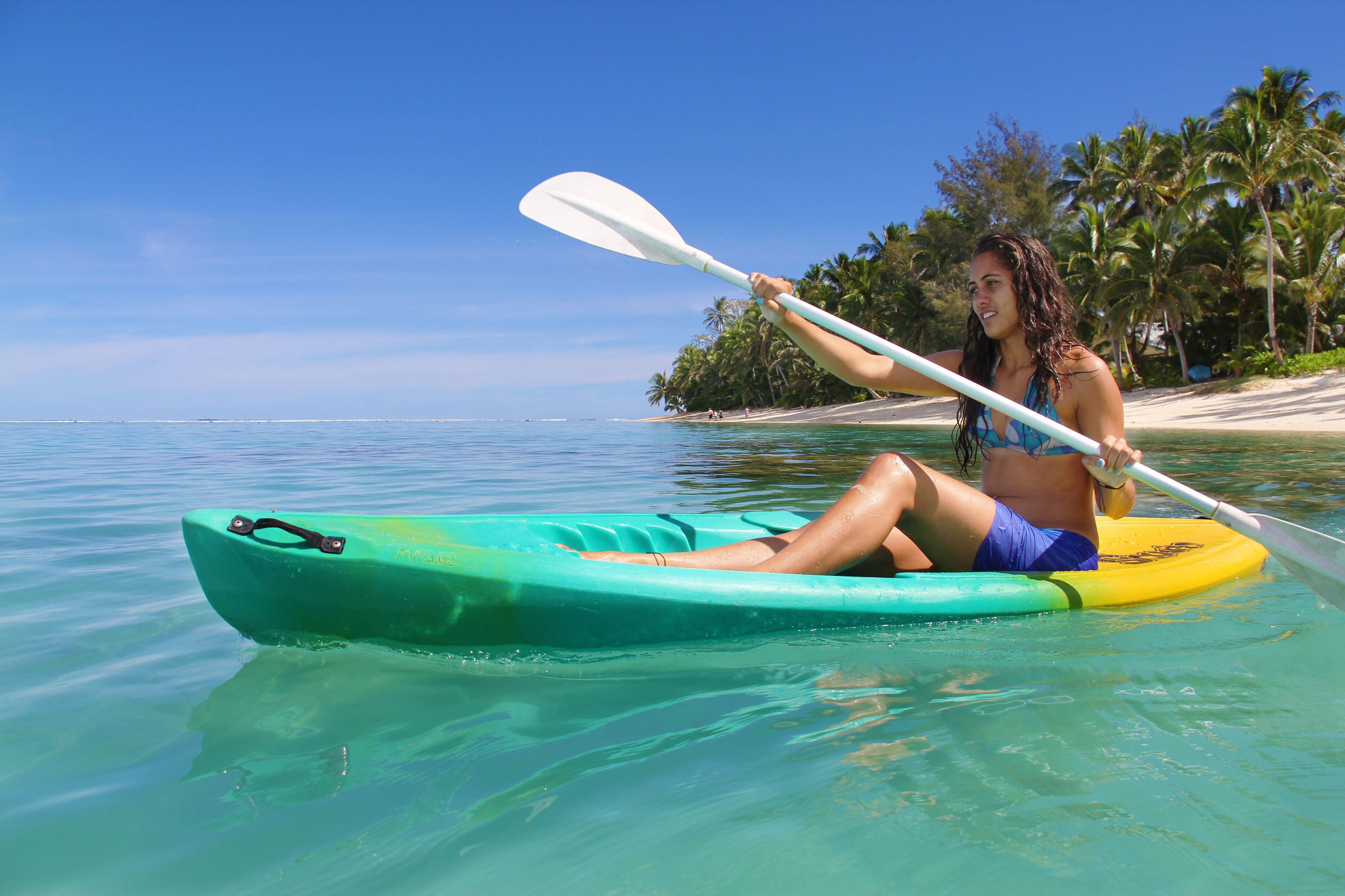A person kayaking on a clear day with a beach and palm trees in the background.