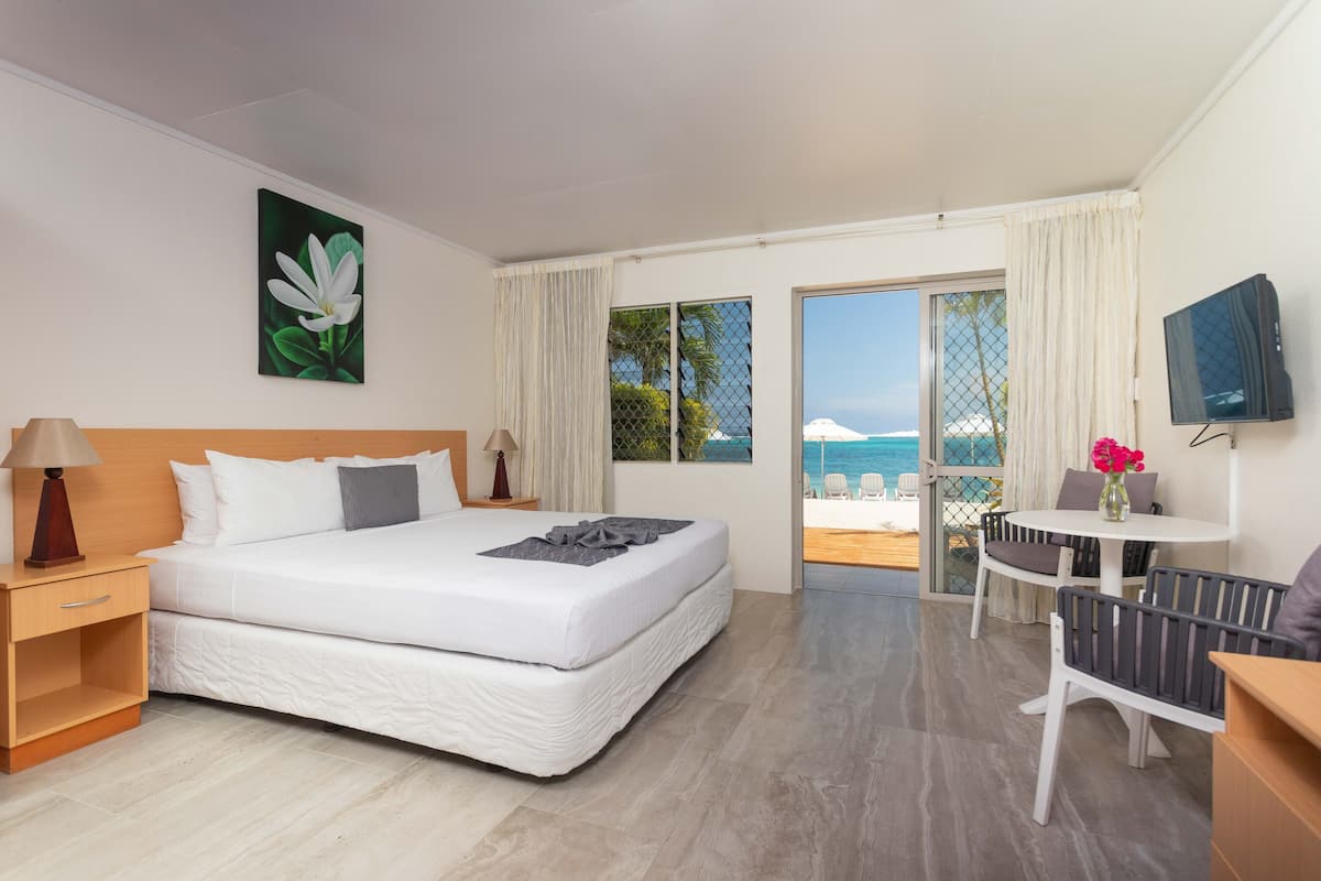 A hotel room with a bed, a TV, a desk with a chair, a small table with a vase of flowers, and a balcony with a view of the sea.