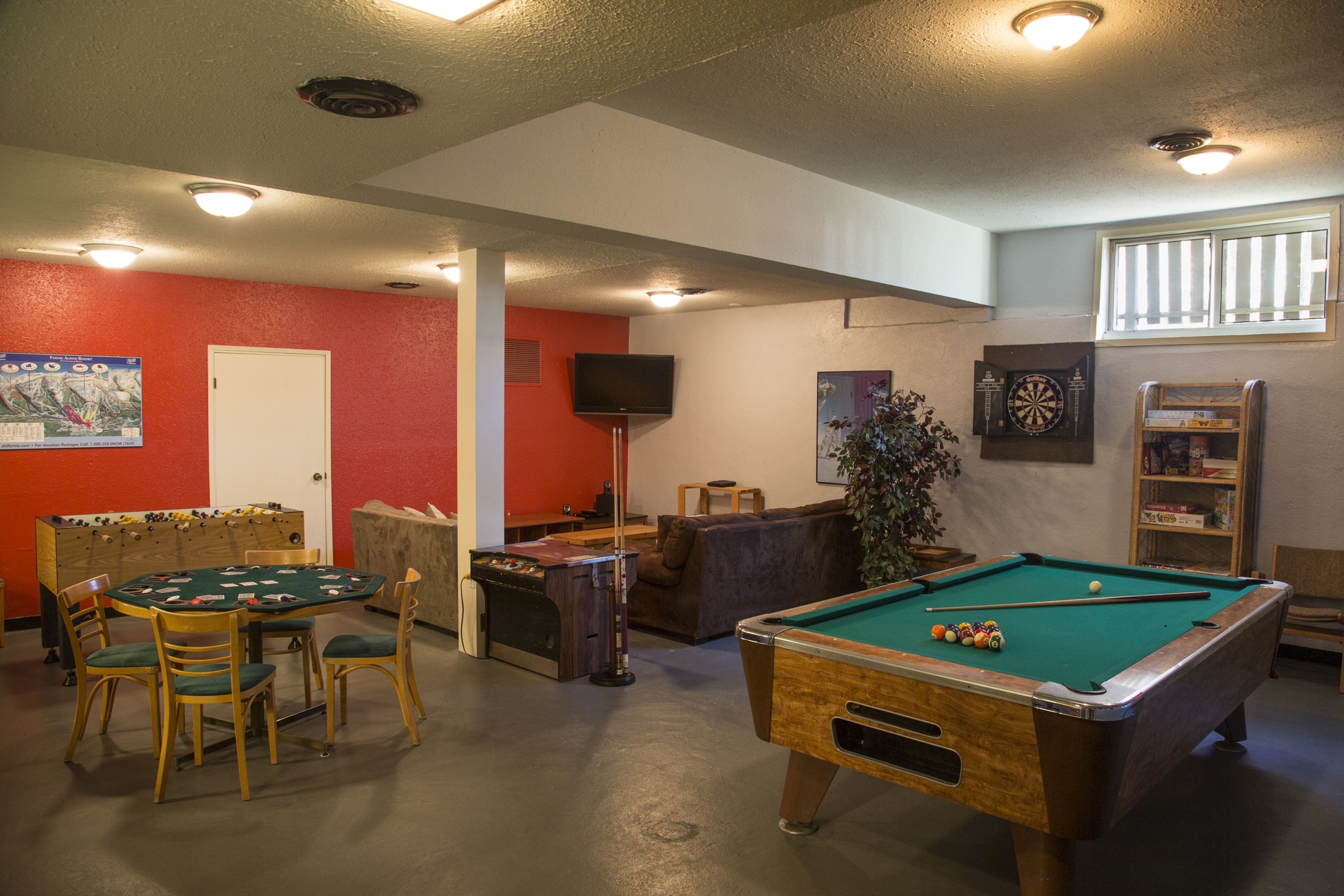 A pool table, a foosball table, and a dartboard in a room with red walls and a window.