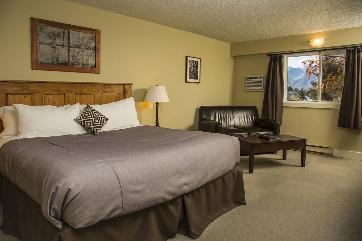 Una habitación de hotel con una cama grande, cabecera de madera, un escritorio, una silla, una lámpara y un ventanal con cortinas.