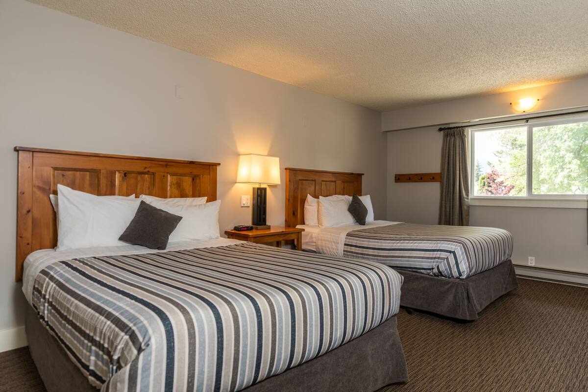 Una habitación de hotel con dos camas, una mesita de noche, una lámpara y un ventanal con cortinas.