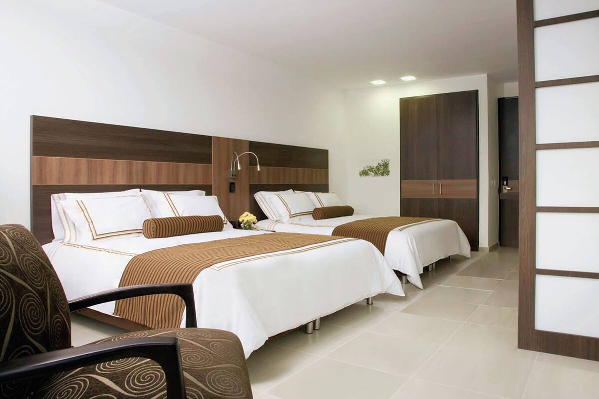 Habitación de hotel con dos camas, cabecera de madera, zona de estar con una silla y una mesita.