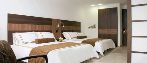 Habitación de hotel con dos camas, cabecera de madera, zona de estar con una silla y una mesita.