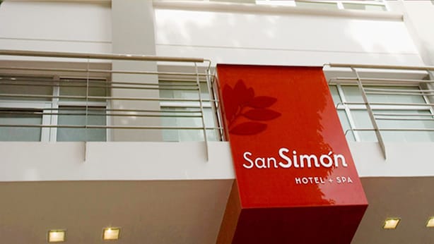 Un letrero rojo con la leyenda 'SanSimon Hotel + Spa', instalado en un edificio con barandilla de vidrio y múltiples ventanales.