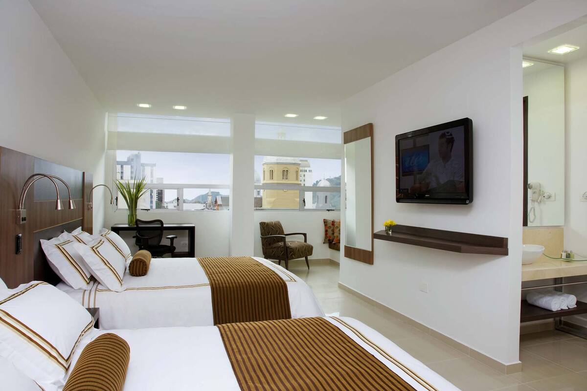 Habitación de hotel con dos camas, un escritorio, una silla, un televisor y un baño con espejo y lavabo.