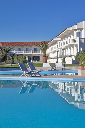 A swimming pool with lounge chairs, a palm tree, and a building with balconies. - Mrs Chryssana Beach Hotel by GHH (Platanias)
