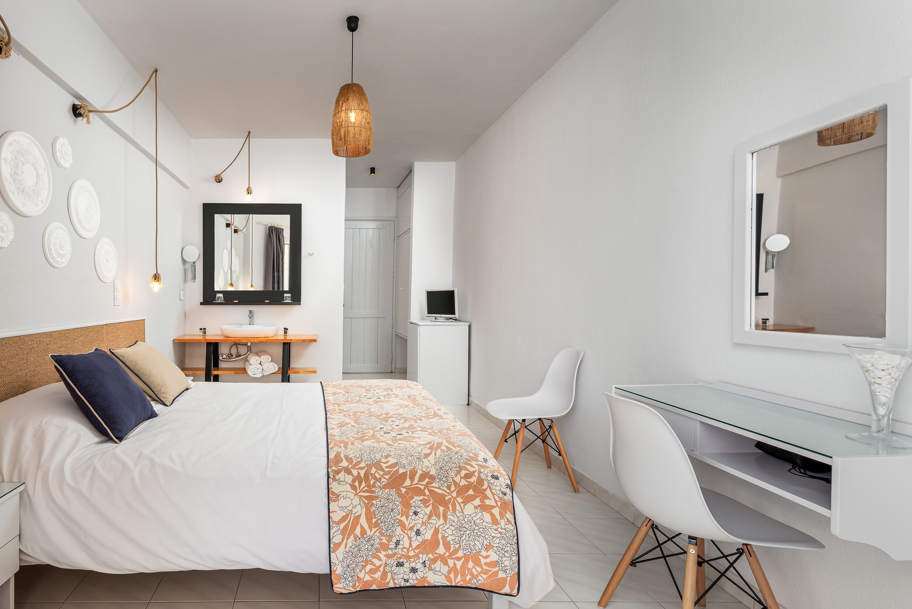 Une chambre moderne avec un lit, un bureau et une chaise, une salle de bain à l’arrière-plan et un luminaire suspendu.