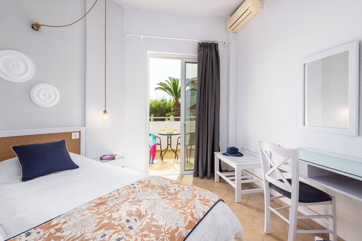 Habitación de hotel con cama, escritorio, silla, balcón con mesa y sillas, y vista a zonas verdes.