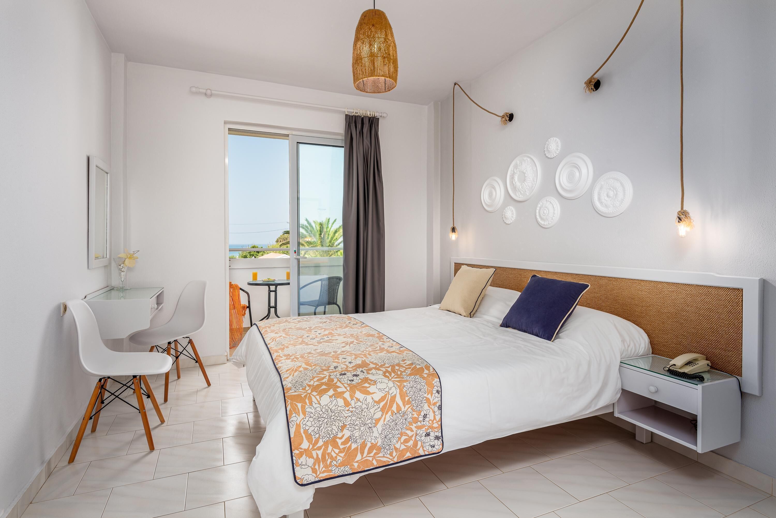 A modern hotel room with a large bed, a desk with chairs, a balcony with a view, and decorative wall plates.