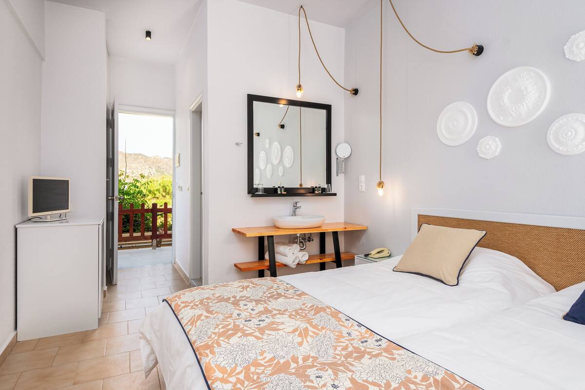 Un dormitorio con cama, mesita de noche, televisor, baño con lavamanos y espejo, y vistas al exterior.