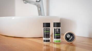 A bathroom sink with two bottles of 'green! tonic' shampoo and shower gel, and a circular soap bar on a wooden surface.