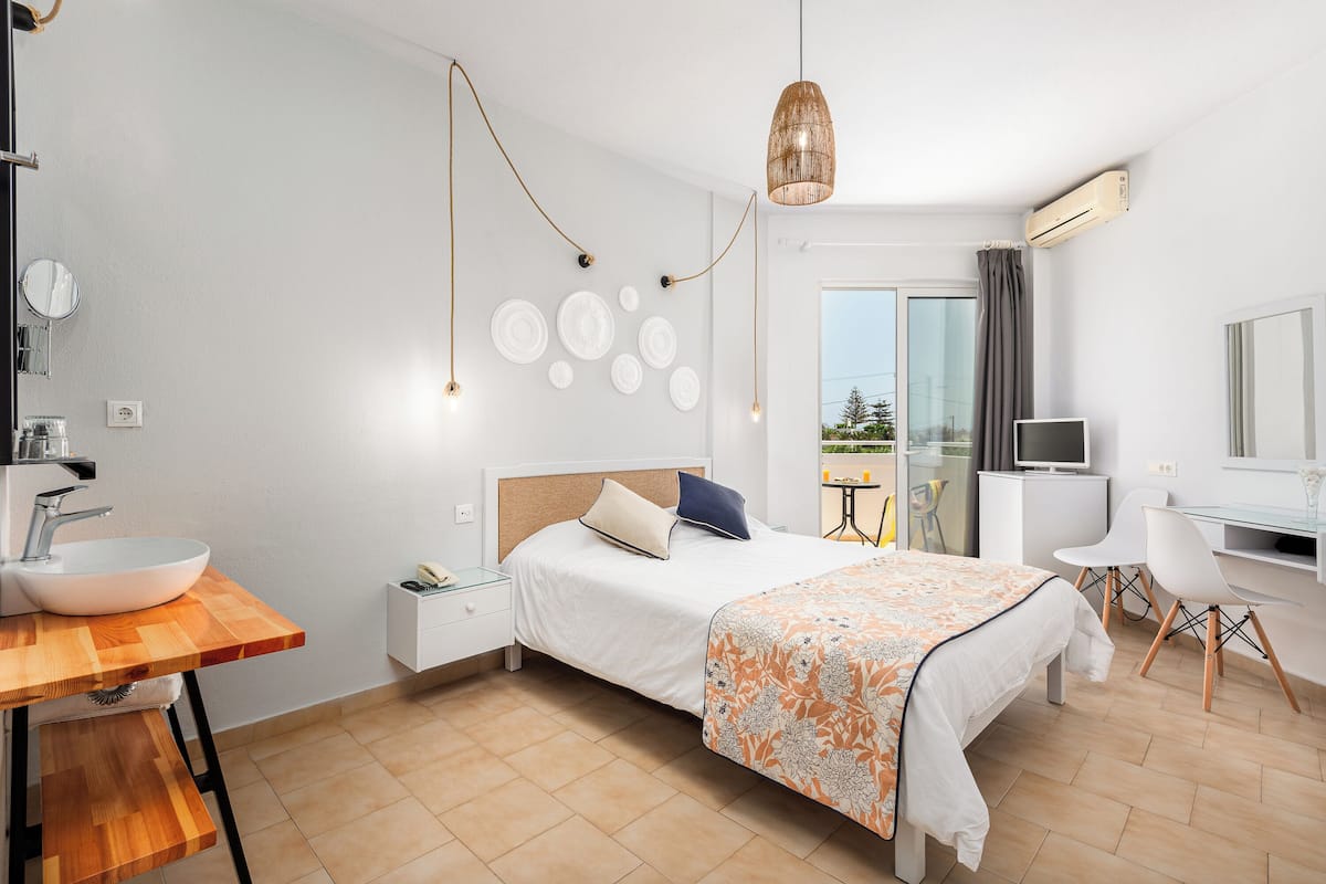 Ein modernes Hotelzimmer mit einem großen Bett, einem Holztisch mit Waschbecken, einem Fernseher und einem Balkon mit Blick.