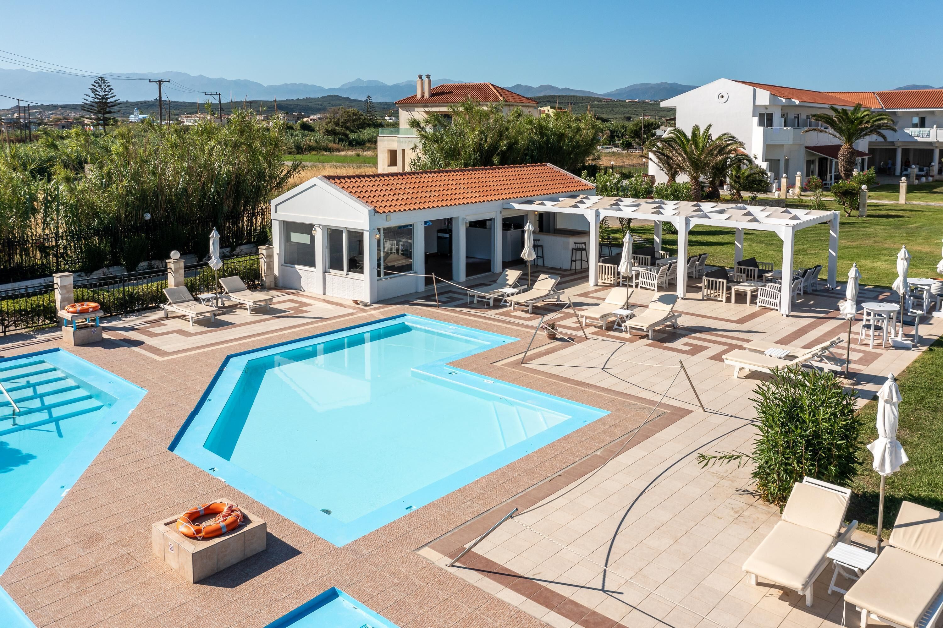 A pool area with multiple sun loungers and a covered seating area, surrounded by a landscaped garden and buildings in the background.