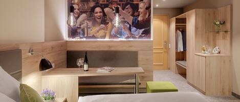 Habitación de hotel moderna con un escritorio de madera, una cama con almohadas verdes, un televisor montado en la pared y un armario con ropa colgada.