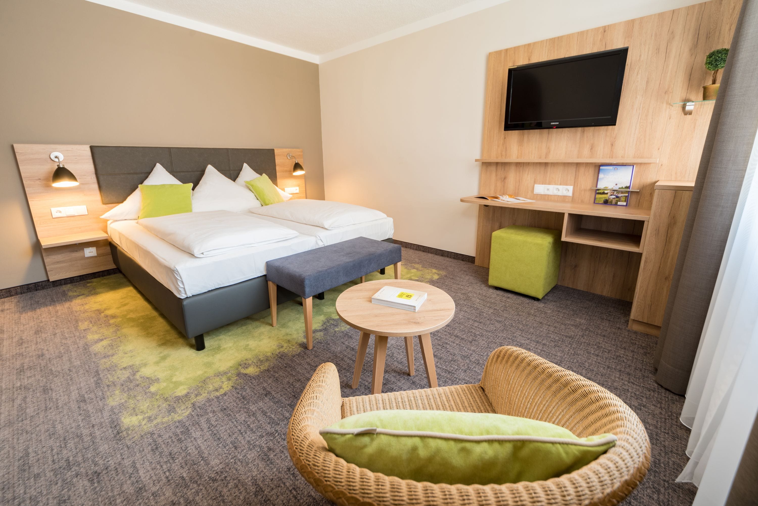 Una habitación de hotel con cama, mesitas de noche, televisor, un pequeño escritorio y una silla de mimbre.